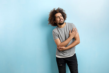 Wall Mural - A curly-headed handsome man wearing a gray T-shirt and ripped jeans is standing and thinking with his hand on the chest, looking upwards over the blue background.