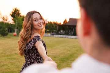 Canvas Print - Joyful young woman holding hand of handsome man while walking outdoor together, on green grass near restaurant or country house. Follow me concept