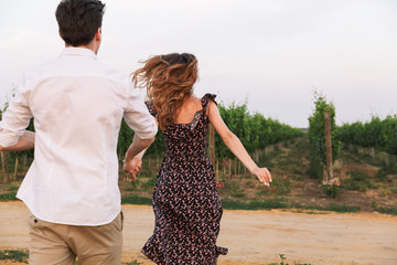 Canvas Print - Photo from back of young caucasian couple man and woman walking outdoor together, through vineyard on summer day