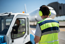 We Doing Great Job. Back View Of Airport Mechanic In Headphones Staring At Colleague. Man In The Car And Airplane On Blurred Background