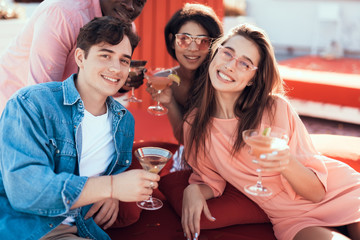 Wall Mural - Satisfied males and beaming women drinking alcohol cocktails while looking at camera. Cheerful comrades relaxing together concept