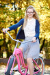 Portrait of blond long-haired attractive traveler girl in glasses, skirt and blouse posing on modern pink lady bicycle on lit by autumn sun park on bright colorful golden bokeh trees background.
