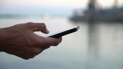Wall Mural - Closeup of a male hand holding a modern phone and surfing the net on a remarkable river embankment with ship refelctions at splendid sunset