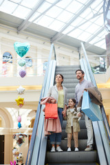 Parents with little girl carrying bags and moving on escalator in light big shopping center enjoying time together