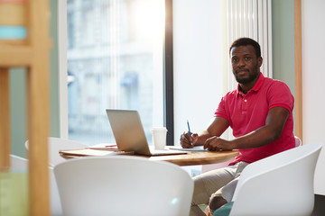 Wall Mural - Adult casual African-American man sitting at table against window with laptop and cup of coffee taking notes and looking at camera