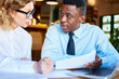© pressmaster - Black man sitting with executive woman at table exploring paper documents and working together
