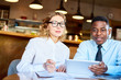 © pressmaster - Serious adult diverse man and woman working together and sitting with tablet and papers at table in cafeteria
