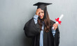 © Krakenimages.com - Young brunette woman over grunge grey wall wearing graduate uniform holding degree stressed with hand on head, shocked with shame and surprise face, angry and frustrated. Fear and upset for mistake.