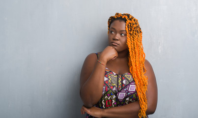 Poster - Young african american woman over grey grunge wall wearing orange braids with hand on chin thinking about question, pensive expression. Smiling with thoughtful face. Doubt concept.