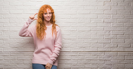 Wall Mural - Young redhead woman standing over brick wall smiling and confident gesturing with hand doing size sign with fingers while looking and the camera. Measure concept.