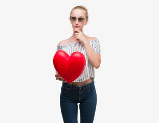 Sticker - Blonde teenager woman holding red heart serious face thinking about question, very confused idea