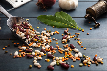 Canvas Print - Close up of assortment of legumes pulses in a stainless scoop, spilled on a wooden tabletop background, pepper grinder, onion, garlic and grater.