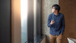 © zephyr_p - Young Asian businessman dressed in casual style using smart phone looking out of window in office building. Urban lifestyle concept