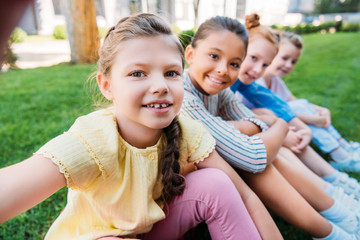 group of happy schoolgirls taking selfie while sitting on grass together