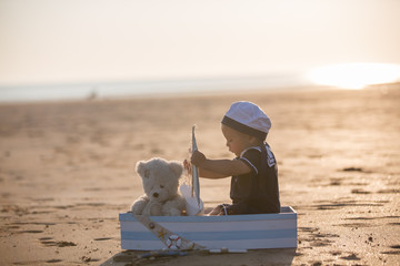 Canvas Print - Cute baby child, sweet boy, playing with boat, teddy bear and fishes on sunset at the edge of the ocean