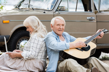 senior wife using laptop while husband playing guitar against beige vintage car