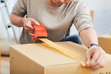 Unrecognizable young man using sticky tape to seal carton box while relocating into new flat