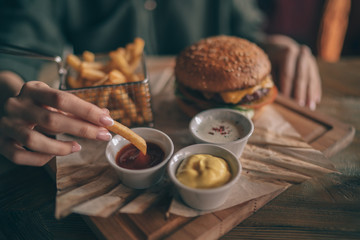 Woman holding cheeseburger and refreshing drink. Young girl holding in hands fast food burger, american meal. Beef burger in hands with french fries on rustic wooden background. Selective focus.