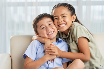 cheerful asian girl smiling and embracing cute boy while sitting on comfortable couch together