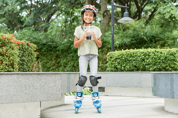 Wall Mural - Cute Asian girl holding cold beverage and smiling while standing on roller skates in park