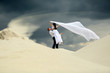 Mężczyzna podnosi wysoko dziewczynę w białej sukni z welonem na piaszczystej wydmie.