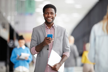 Wall Mural - Cheerful adult African-American man standing with smartphone and papers and looking at camera in office hall