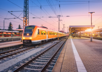 Passenger high speed train on the railway station at sunset. Urban landscape with modern commuter train on the railway platform against colorful sky at dusk. Intercity vehicle on railroad in Europe