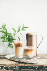 Poster - Iced coffee in glasses with milk and straws on board over rustic wooden table, white wall, jug and plant branch in vase at background, copy space. Summer refreshing beverage, ice coffee drink concept