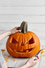 Woman carving big orange pumpkin into jack-o-lantern for Halloween holiday decoration on white wooden planks, close up view