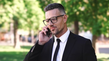 Poster - Happy businessman dressed in shirt talking by smartphone while standing in park