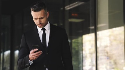 Wall Mural - serious business man dressed in suit using smartphone while waitng someone outside a glass building