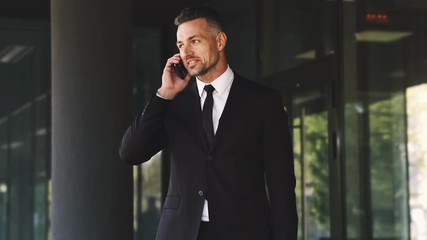 Wall Mural - smiling business man dressed in suit talking by smartphone while standing outside a glass building