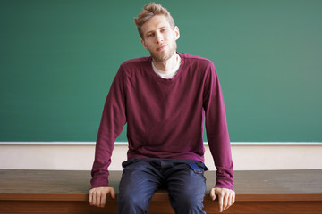 young bearded student sits on the teacher table desk in front of blackboard with copy space