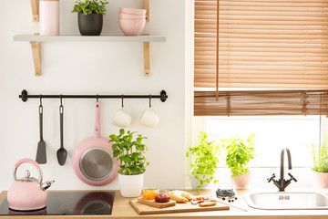 Real photo of wooden countertop with fresh plants, board with croissant, marmalades and fruits and pink kitchen tools