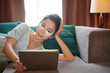 © DragonImages - Pretty Asian female with under-eye patches smiling and browsing tablet while lying on comfortable couch in living room
