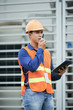 © DragonImages - Attractive Asian man in hardhat and waistcoat looking away and talking on walkie-talkie while standing on construction site
