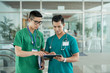 © DragonImages - Two Asian health professionals in medical scrubs standing in hospital hall and browsing tablet together