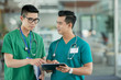 © DragonImages - Two young Asian men in medical scrubs pointing at information on tablet screen while standing on blurred background of hospital hall