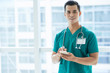 © DragonImages - Attractive Asian man in medical uniform smiling and looking at camera while standing near hospital window and using smartphone