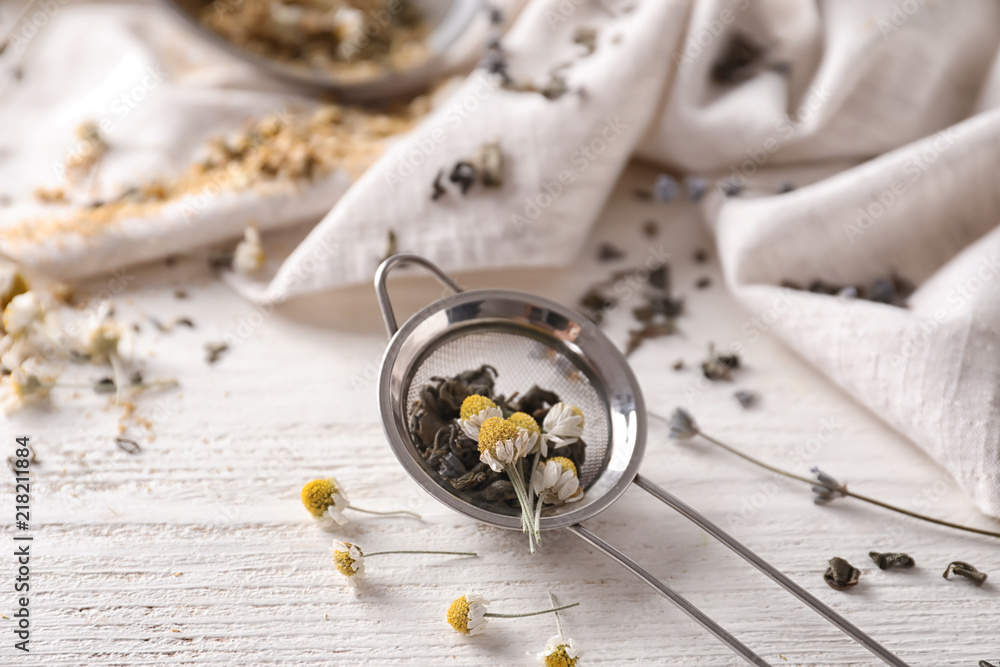 Sieve with dried chamomile and tea leaves on white wooden table