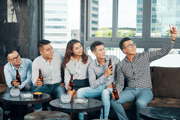 Wall Mural - Relaxing cheerful Asian people sitting in bar and taking selfie with smartphone while having beer