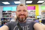 Fototapeta  - Bearded man makes a selfie portrait in a gadget store and computer equipment.