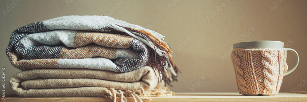 stack of beige checkered wool blankets and cup on a wooden chest. autumn - winter concept of home warmth and comfort. toned, long banner - obrazy, fototapety, plakaty 