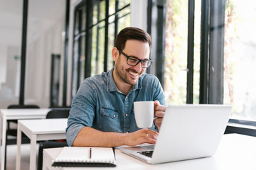 Wall Mural - Handsome freelancer using laptop and drinking coffee in bright modern enviroment.