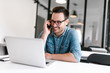 © bnenin - Smiling young man talking on smartphone while sitting in front of laptop.