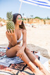 © F8  \ Suport Ukraine - Close up summer portrait of stylish joyful happy sexy woman with perfect tanned fit body wearing trendy sunglasses, holding a pineapple, tropical fruit lying under umbrella at the beach