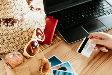 booking summer vacation trip online. traveling holidays and tourism concept. woman holding credit or debit bank card paying for the tour on the internet. laptop hat sunglasses on the desk.