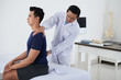 © DragonImages - Osteopath checking spine of sitting young Vietnamese man