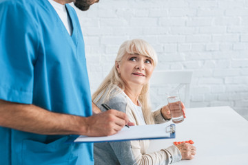 cropped shot of caregiver writing on clipboard while senior woman holding glass of water and pills