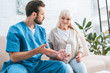 © LIGHTFIELD STUDIOS - young male social worker showing medicine to senior woman sitting on couch
